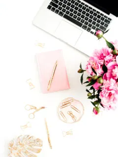 A workspace with laptop, pink peonies bouquet, golden accessories, pink diary on white background.